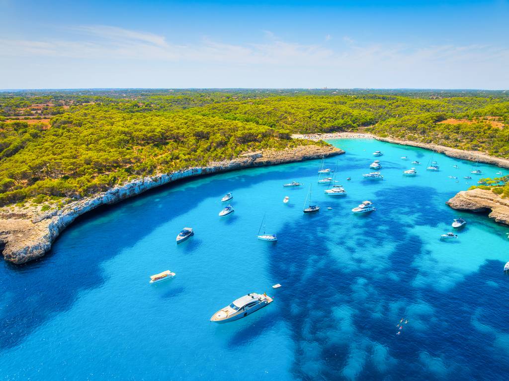 Veduta aereadi Cala Mondragó, una baia turchese con barche ancorate vicino a una riva boscosa e verde, sotto un cielo azzurro limpido.
