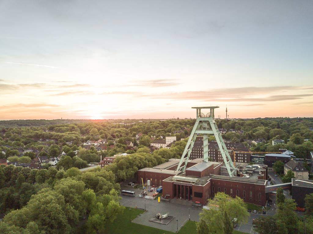 Luftaufnahme eines Förderturms, umgeben von Bäumen und Gebäuden bei Sonnenuntergang, mit klarem Himmel und der Sonne nahe dem Horizont