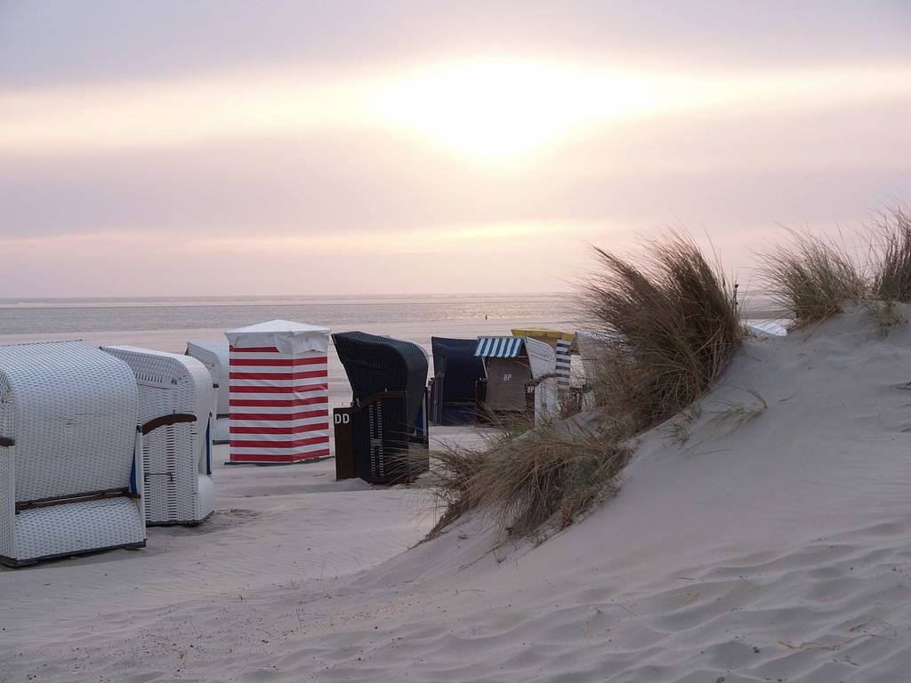 Reihe gestreifter Strandkörbe entlang von Sanddünen mit hohem Strandgras bei einem pastellfarbenen Sonnenuntergang.
