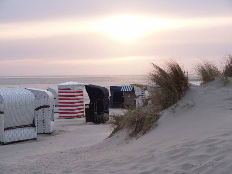 Reihe gestreifter Strandkörbe entlang von Sanddünen mit hohem Strandgras bei einem pastellfarbenen Sonnenuntergang.