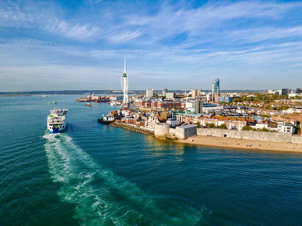Aerial view of a ferry sailing near a coastal city with a prominent white tower and waterfront buildings under a clear blue sky.