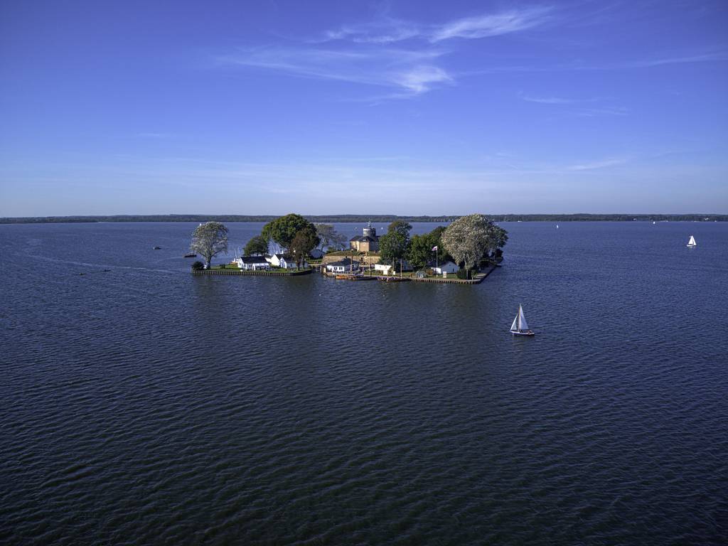Eine kleine, bewaldete Insel mit Gebäuden, umgeben von einem weiten blauen See unter klarem Himmel; in der Nähe segelt ein Segelboot.