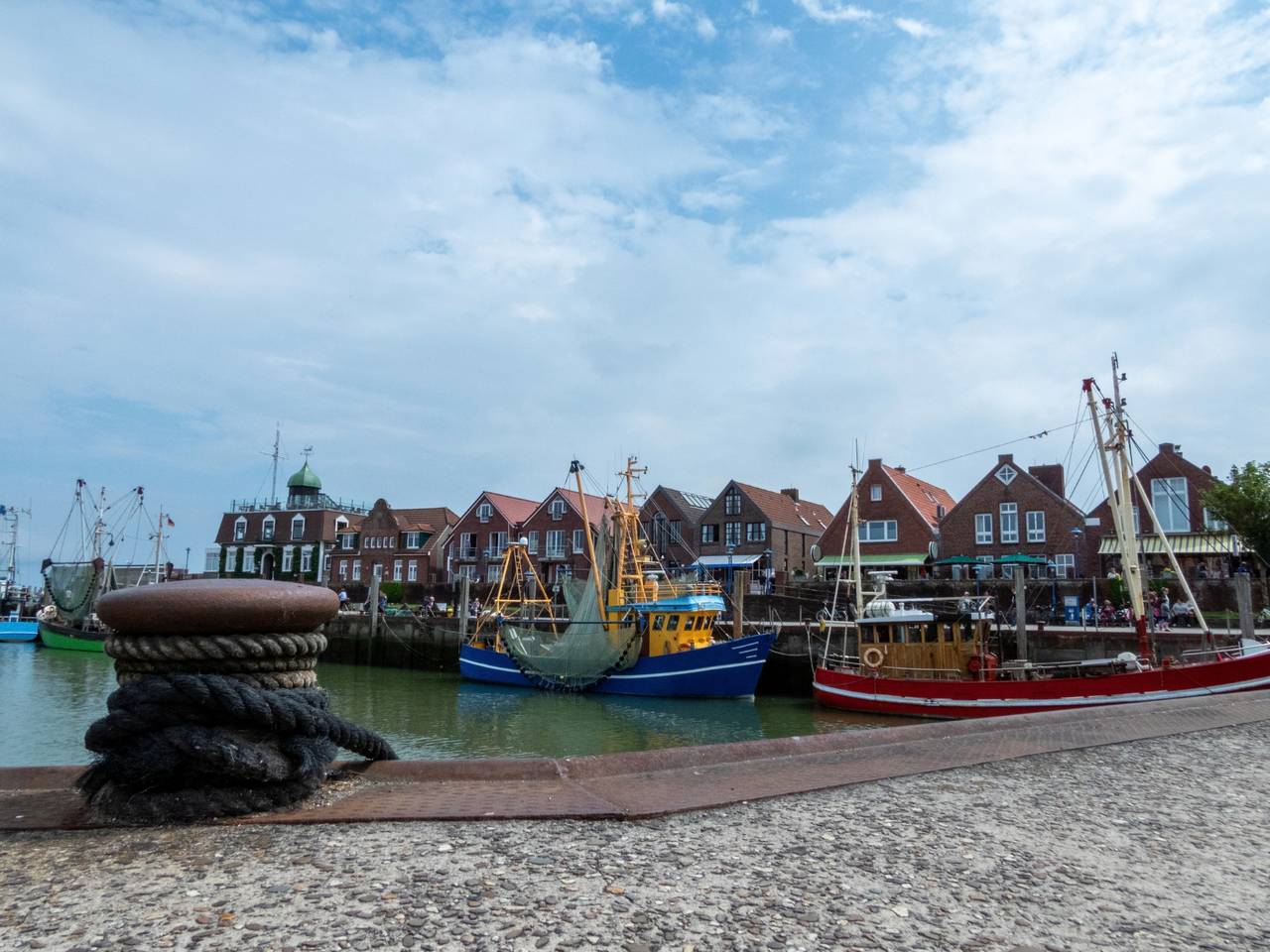 Angedockte Boote in einem malerischen, von Backsteingebäuden gesäumten Hafen unter bewölktem Himmel. Im Vordergrund ist ein aufgerolltes Seil zu sehen.