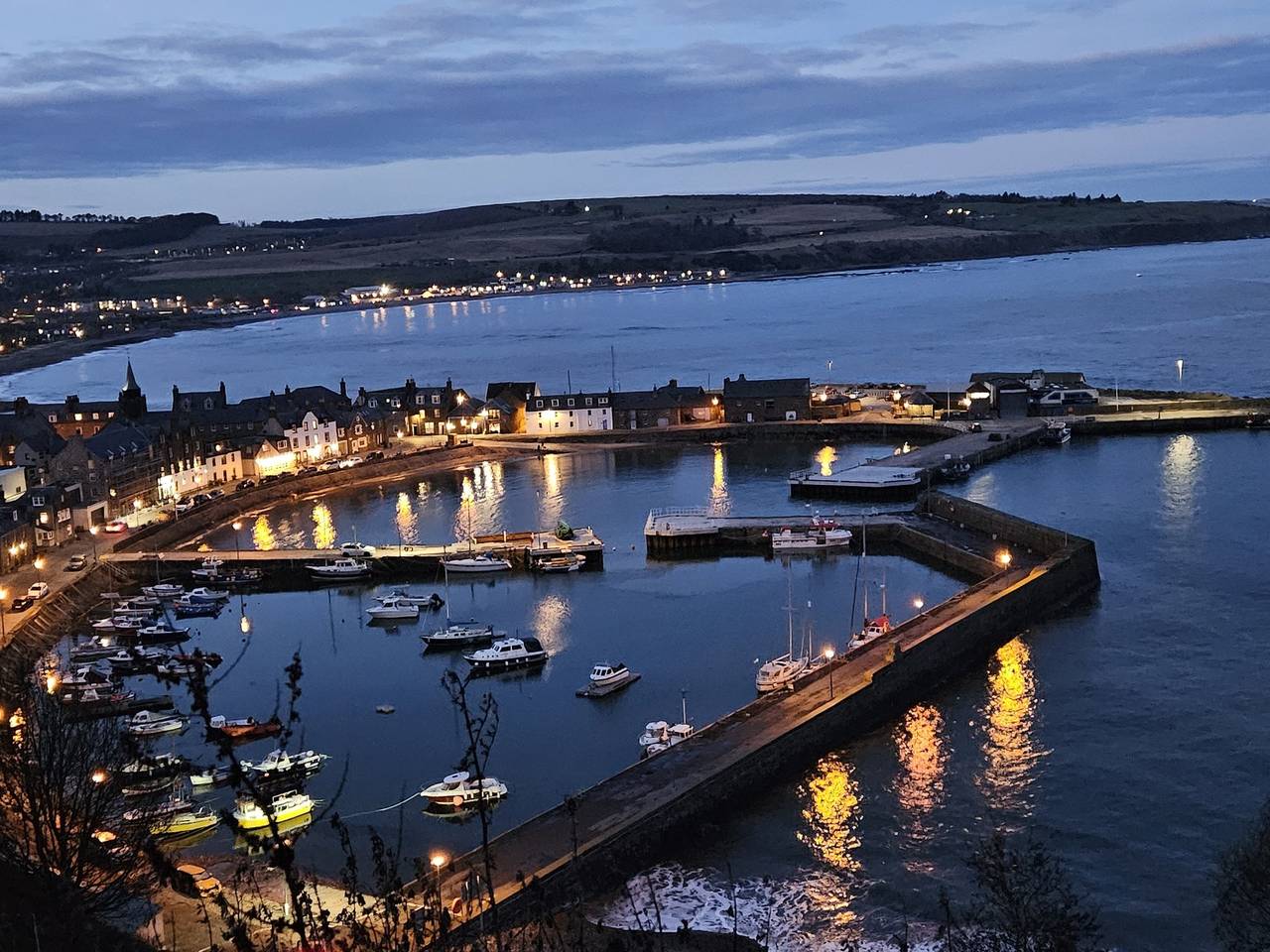 Scenic view of a coastal harbor at twilight with boats docked, surrounded by lit up buildings and calm water reflecting lights.