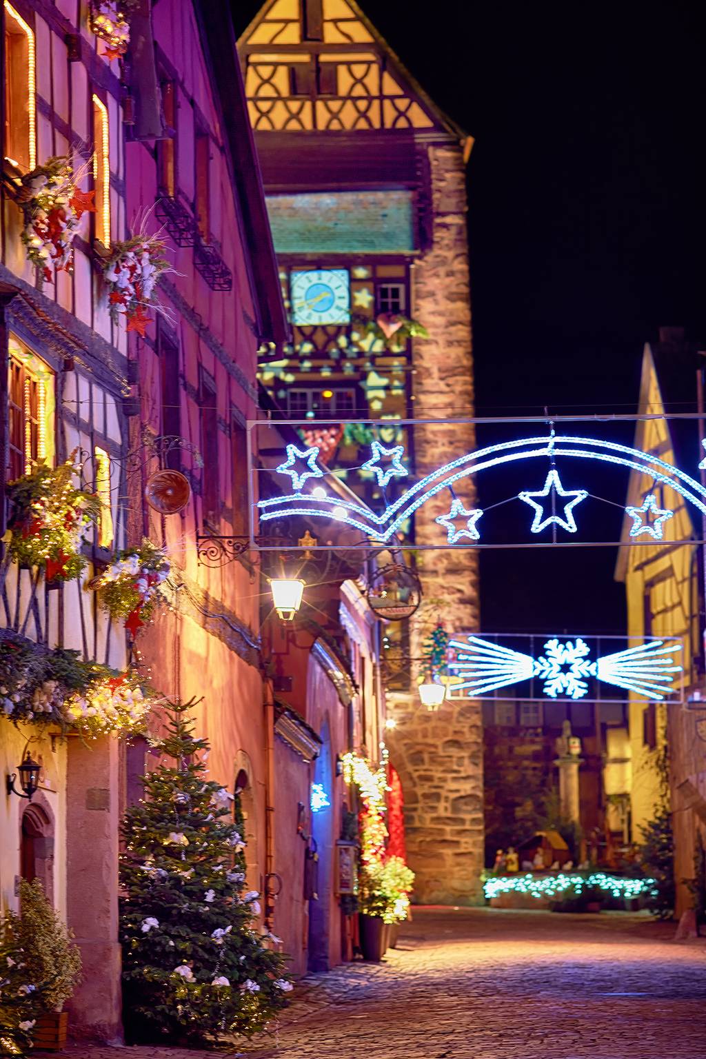 Eine idyllische Kopfsteinpflasterstraße mit festlicher Weihnachtsdekoration, die nachts von bunten Lichtern und einem markant beleuchteten Uhrturm erhellt wird.