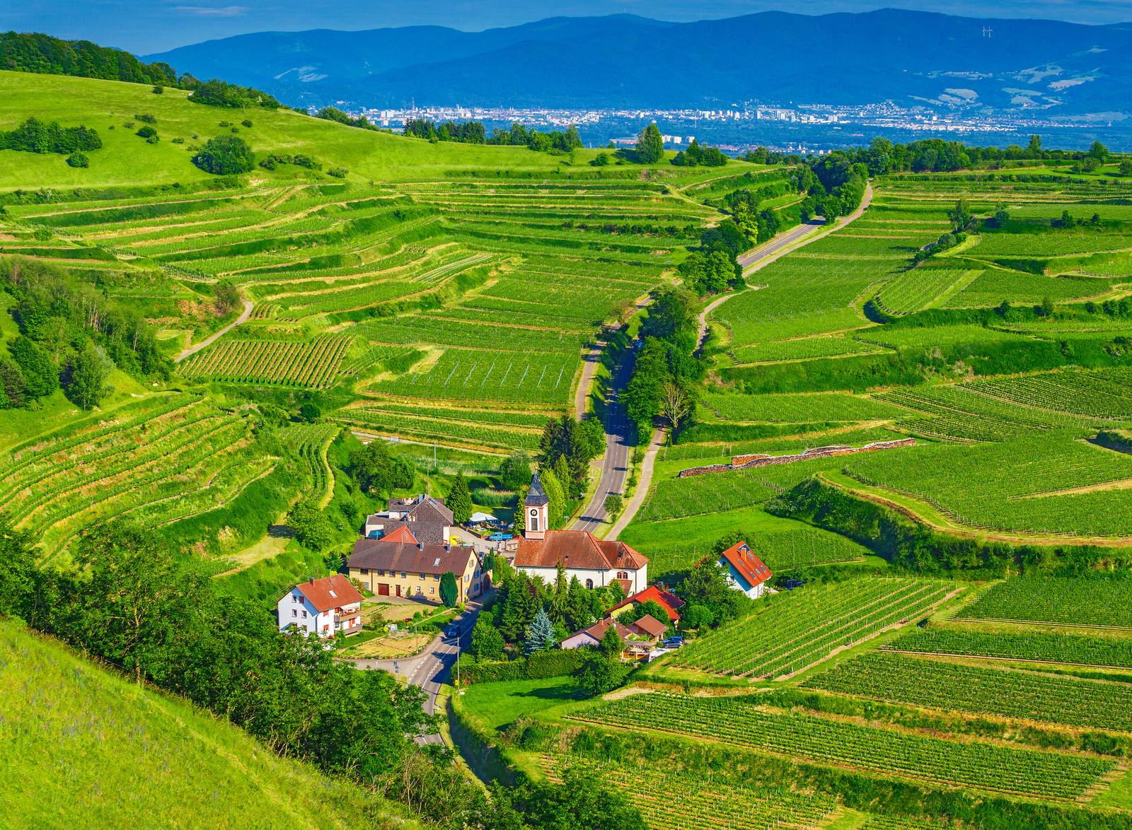 Blick auf ein Dorf, umgeben von grünen Terrassenfeldern und Hügeln, mit einer fernen Stadt und Bergen im Hintergrund