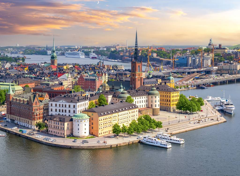 Aerial view of a historic European city by the water, featuring colorful buildings, a church with a tall spire, and several boats docked along the waterfront.