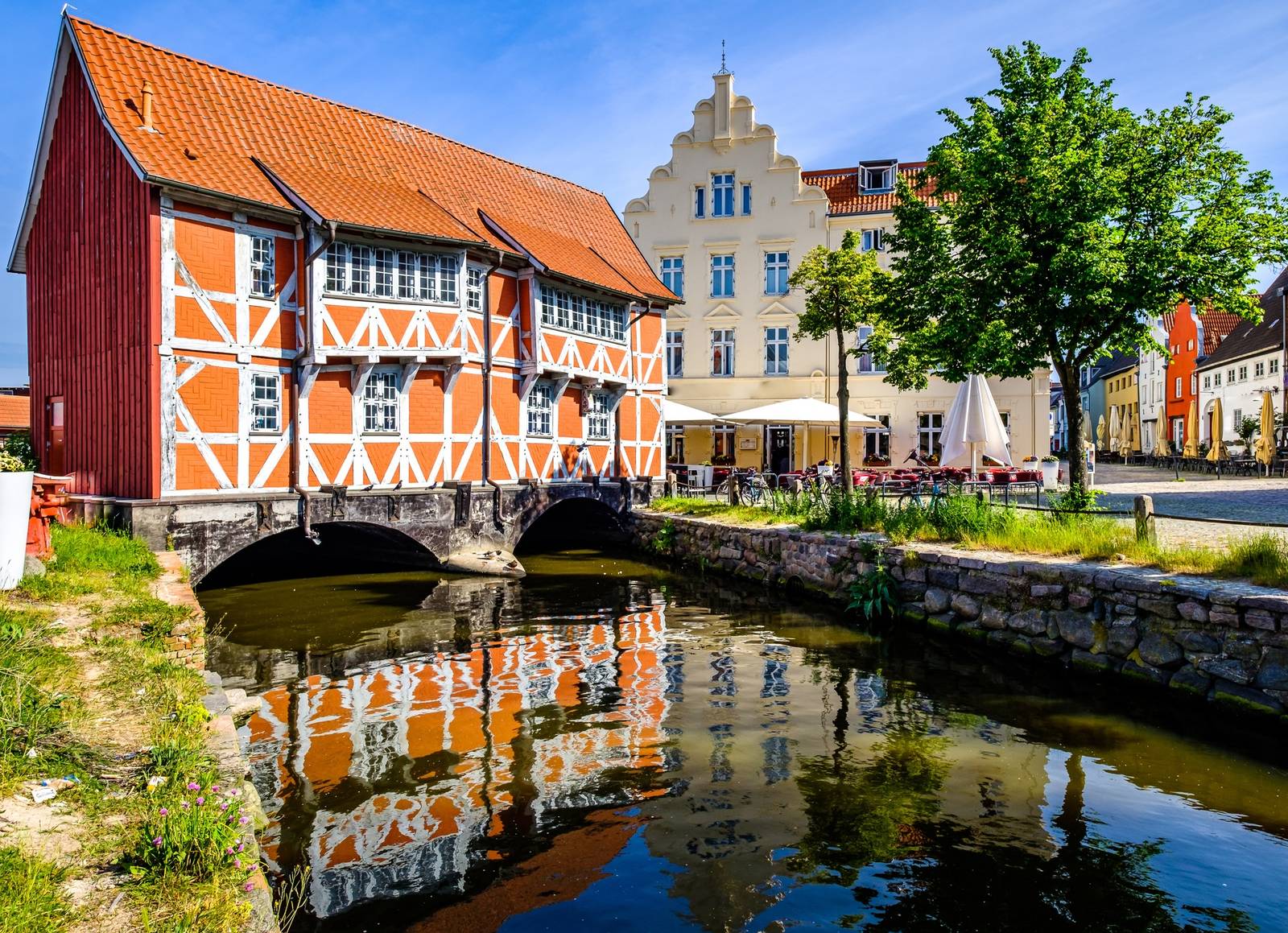 Historisches Fachwerkgebäude mit rotem Dach und Spiegelung im Kanal, neben einem giebelständigen cremefarbenen Gebäude, einem Baum und Außensitzplätzen unter blauem Himmel.