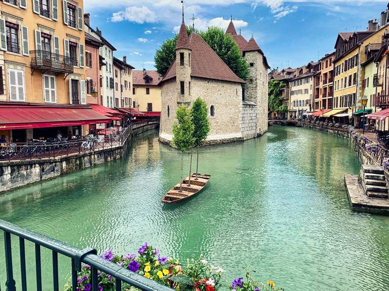 Ein malerischer Kanal mit einem kleinen Boot, umgeben von bunten Gebäuden und Blumen, unter einem teilweise bewölkten Himmel.