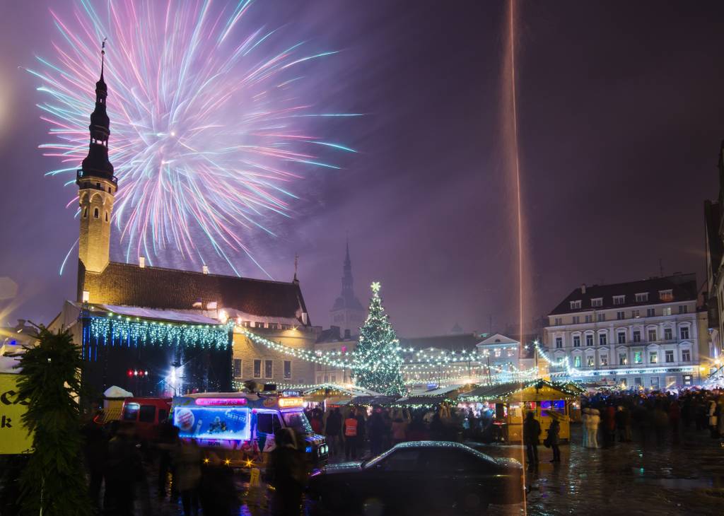 Festive nighttime scene with fireworks over a town square, a lit Christmas tree, market stalls, and a church with a tall spire.