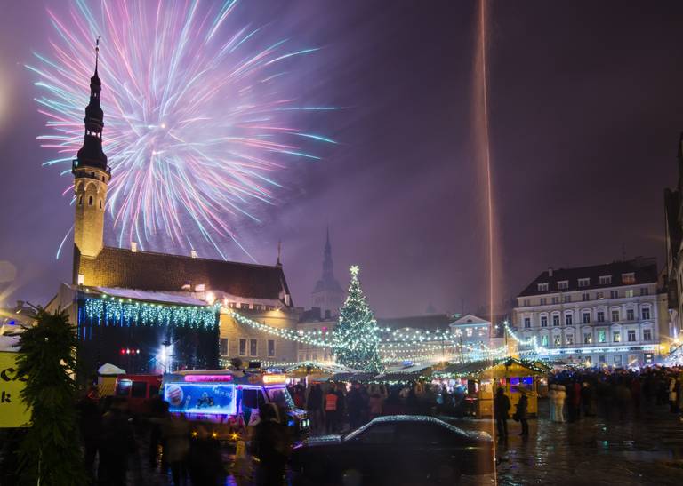 Festive nighttime scene with fireworks over a town square, a lit Christmas tree, market stalls, and a church with a tall spire.
