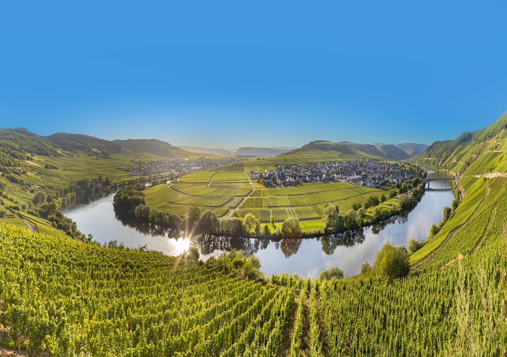 „Panoramablick auf einen Fluss, der sich durch grüne Weinberge und eine Kleinstadt windet, unter blauem Himmel