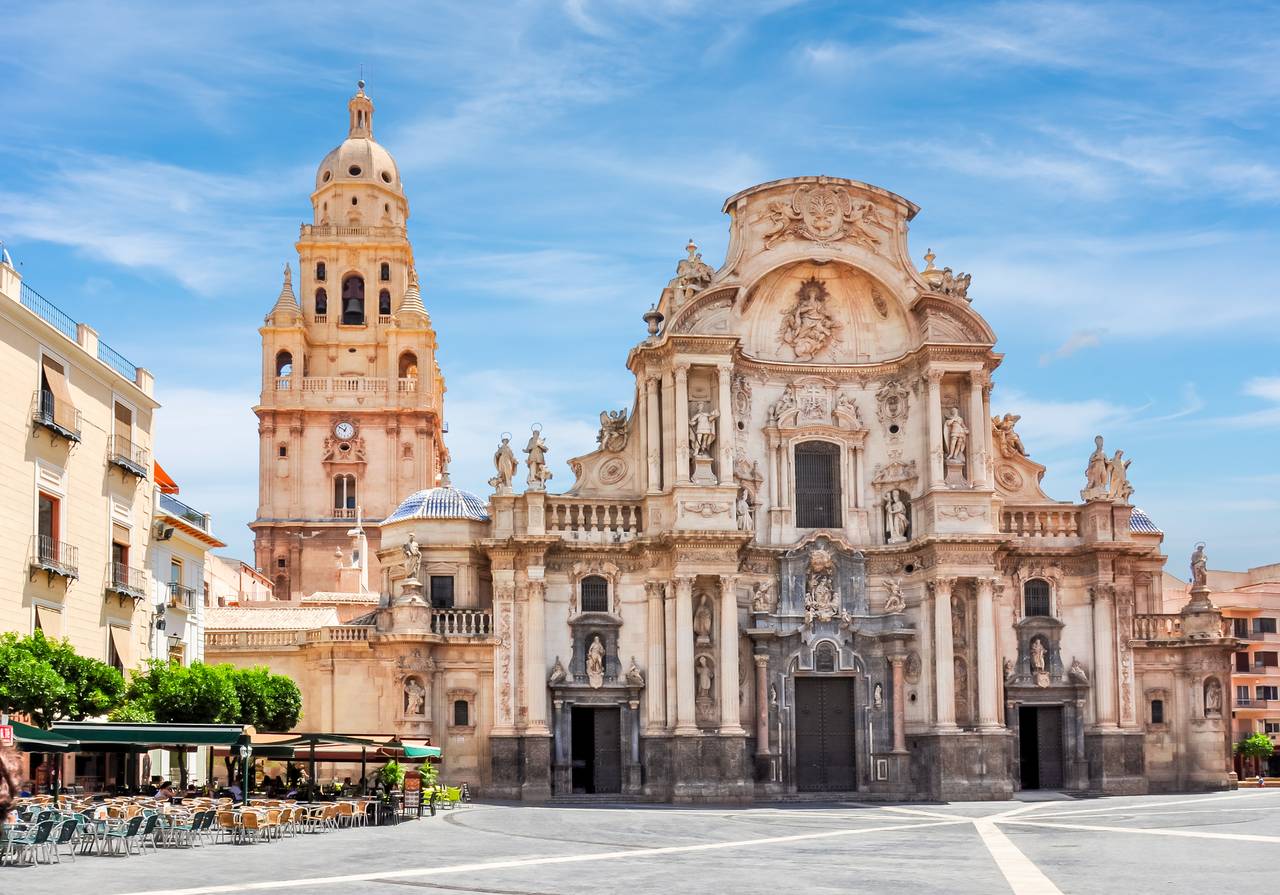 Catedral barroca ornamentada con un alto campanario en una plaza soleada, rodeada de edificios históricos y terrazas de cafeterías al aire libre.