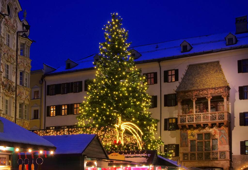 Ein großer, mit Lichtern geschmückter Weihnachtsbaum steht auf einem festlich beleuchteten Marktplatz, umgeben von historischen Gebäuden unter einem tiefblauen Abendhimmel.