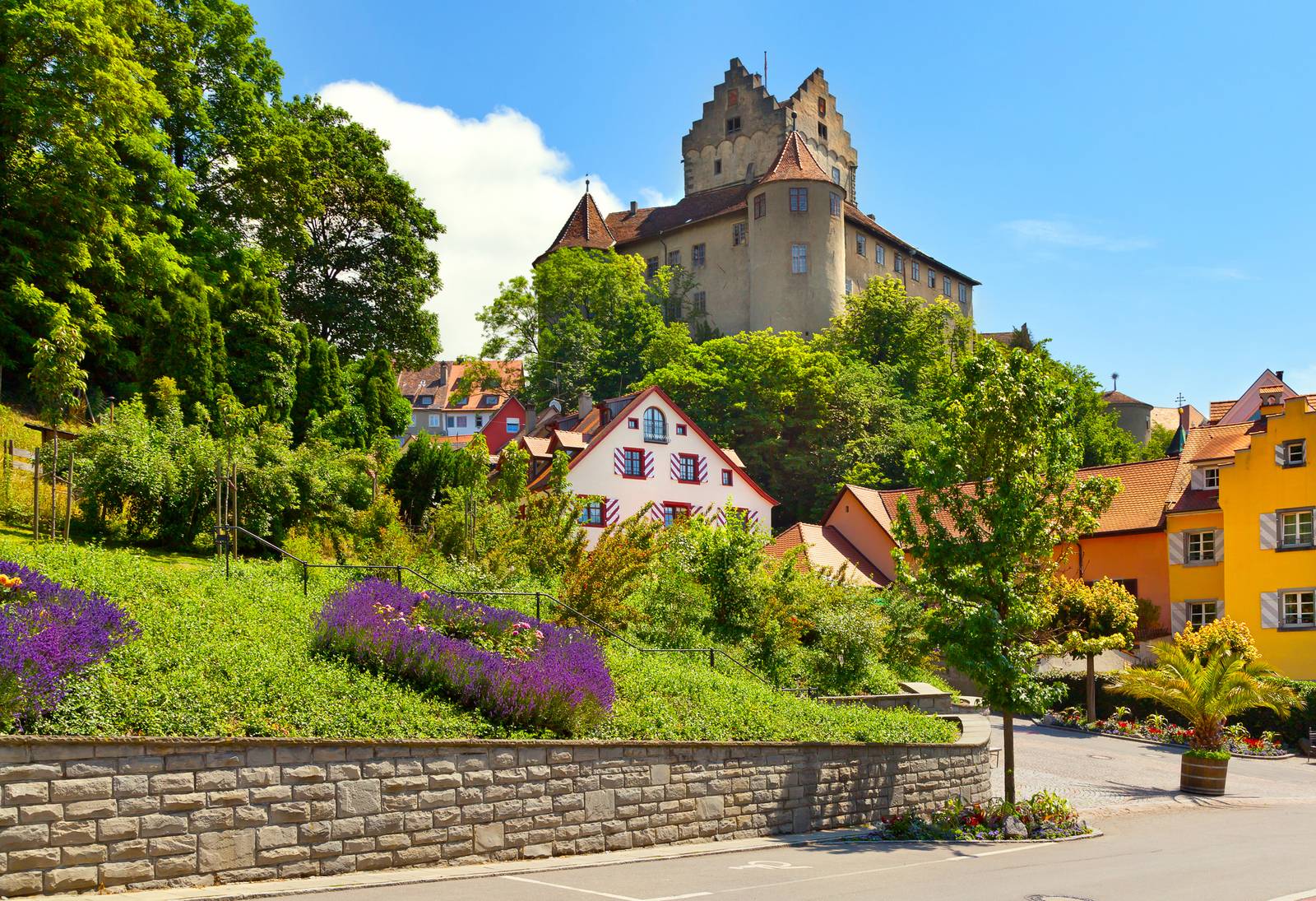 Historisches Schloss auf einem Hügel mit farbenfrohen Häusern und üppigem Grün im Vordergrund unter klarem, blauem Himmel