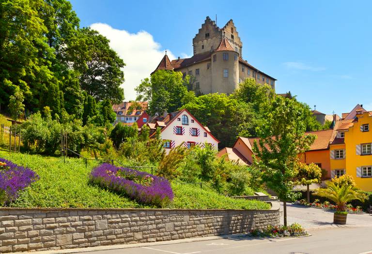 Historisches Schloss auf einem Hügel mit farbenfrohen Häusern und üppigem Grün im Vordergrund unter klarem, blauem Himmel
