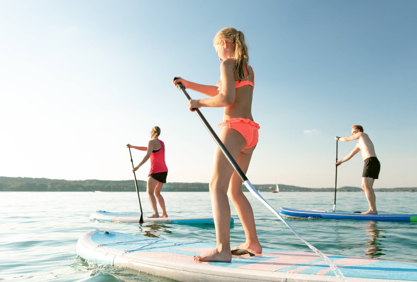 Drei Personen beim Paddleboarding auf einem ruhigen See unter klarem blauem Himmel, gekleidet in Badebekleidung und Schwimmwesten.