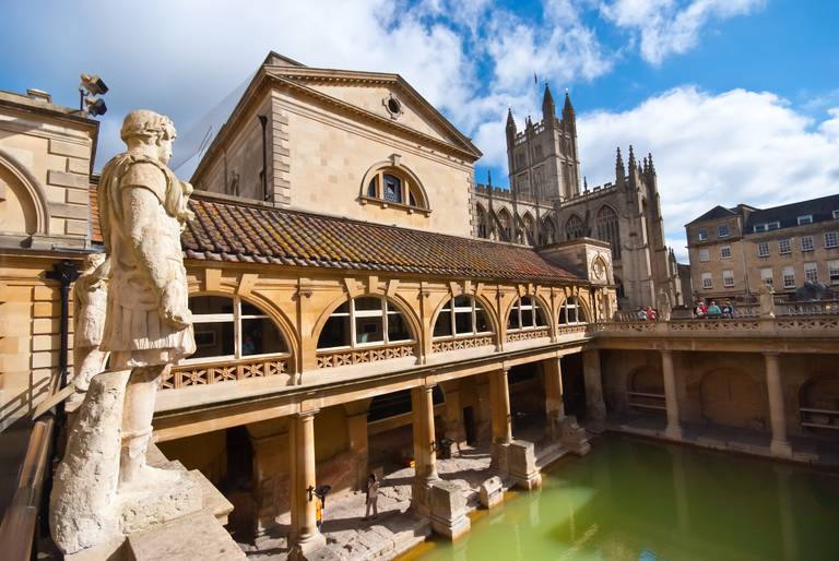 Les thermes romains de Bath, en Angleterre, avec une architecture historique, une statue et un bassin vert sous un ciel bleu avec des nuages.