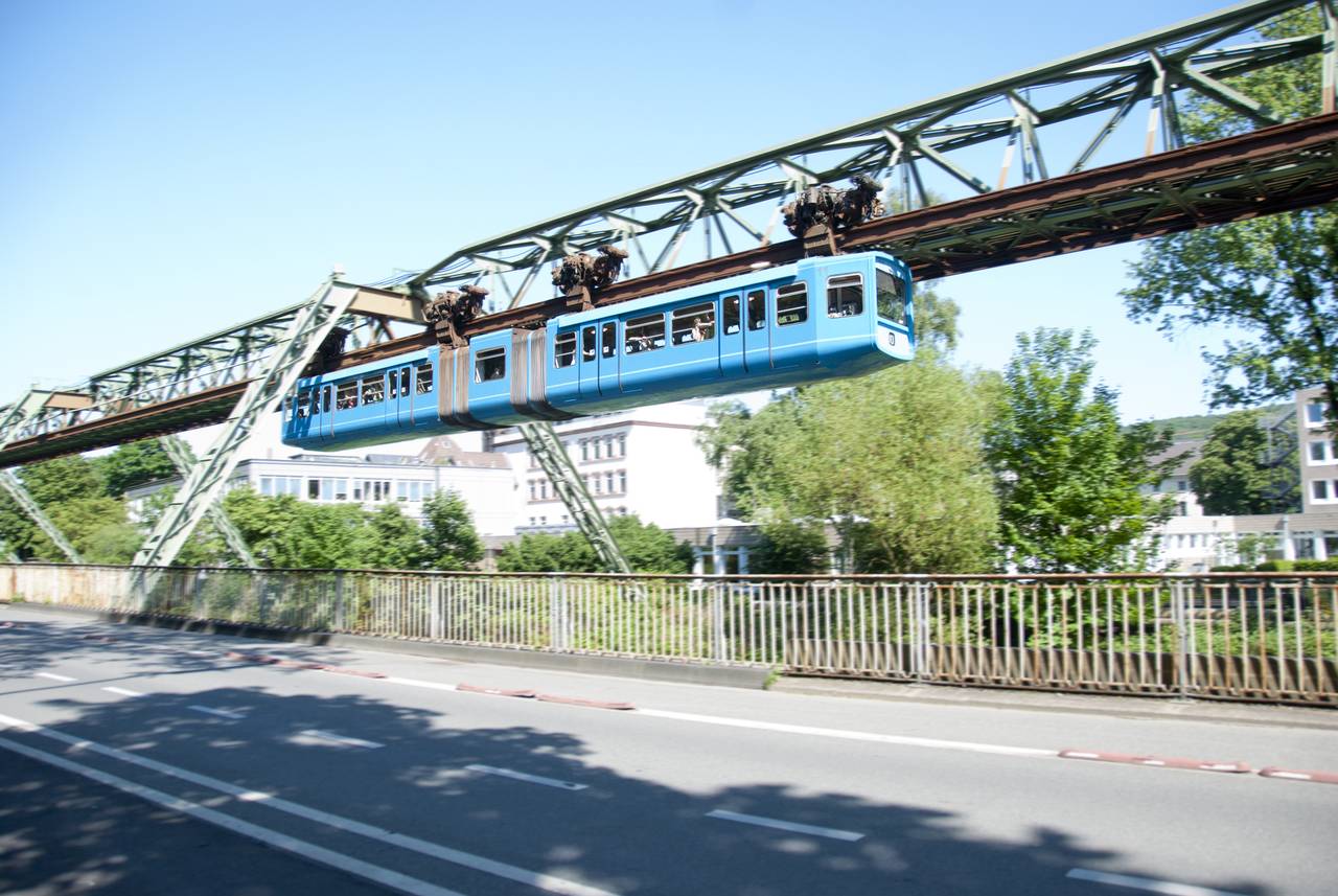 Eine blaue Schwebebahn fährt an einem klaren Tag über eine Straße, im Hintergrund Bäume und Gebäude.