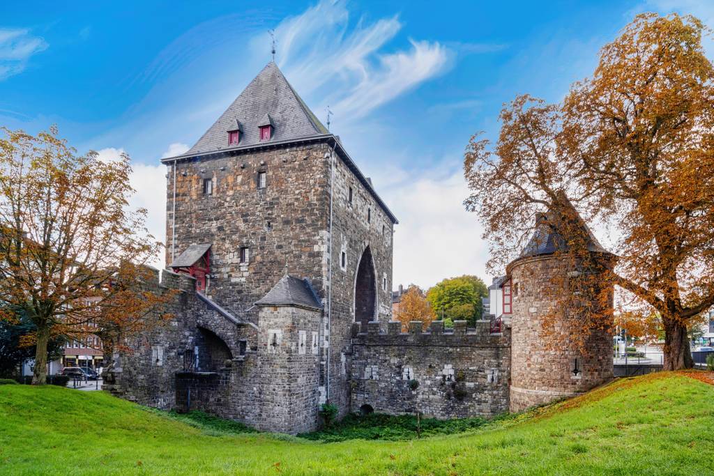 Medieval stone gatehouse with towers, autumn trees and green lawn under a bright blue sky.