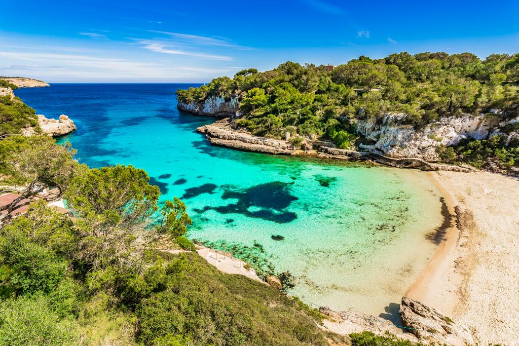 Veduta panoramica di Cala Llombards , una baia turchese dalle acque cristalline, circondata da scogliere rocciose e vegetazione lussureggiante sotto un cielo azzurro limpido, con una spiaggia di sabbia in primo piano.