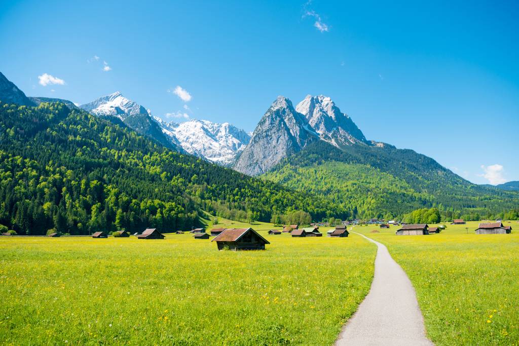 Ein Weg führt durch eine leuchtend grüne Wiese, die mit kleinen Hütten übersät ist, vor der Kulisse schneebedeckter Berge und eines klaren blauen Himmels.