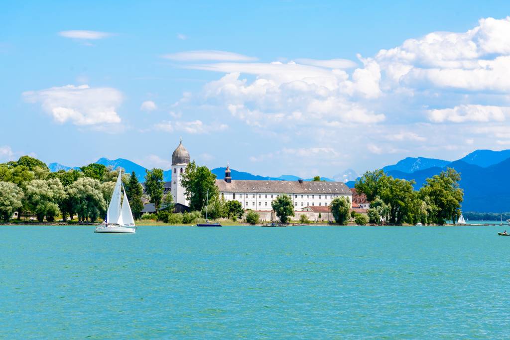 Ein Segelboot gleitet über einen ruhigen See, während am Ufer ein historisches Gebäude von üppigen Bäumen umgeben ist, unter einem hellen, bewölkten Himmel.
