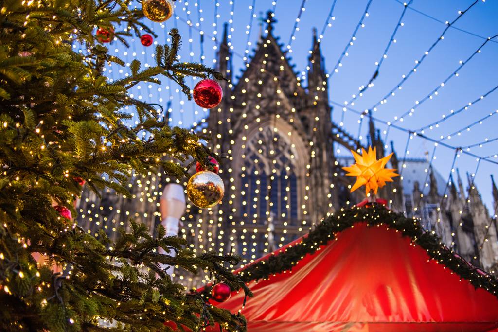 Een kerstmarkt met een versierde kerstboom, feestelijke verlichting en een rode tent op de voorgrond, met op de achtergrond een gotische kathedraal.