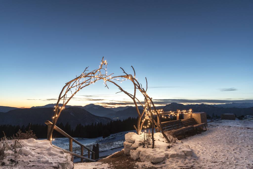 Verschneite Berglandschaft bei Sonnenuntergang mit einem aus Ästen gefertigten Bogen, der mit Lichterketten geschmückt ist, und Sitzbänken in der Nähe.