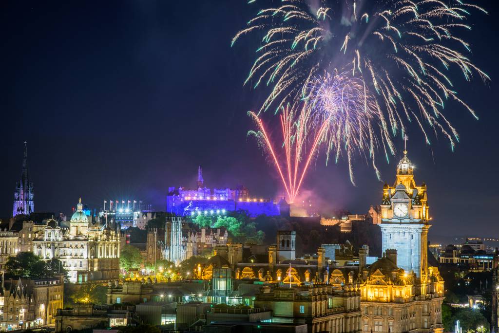 Fireworks illuminate the night sky above a lit-up Edinburgh Castle, with surrounding historic buildings aglow in warm lights.