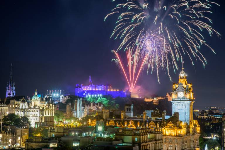 Fireworks illuminate the night sky above a lit-up Edinburgh Castle, with surrounding historic buildings aglow in warm lights.