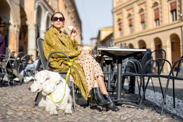 Eine Frau mit Sonnenbrille und grünem Tuch trinkt einen Espresso in einem gepflasterten Straßencafé, während ihr weißer Hund angeleint zu ihren Füßen ruht.