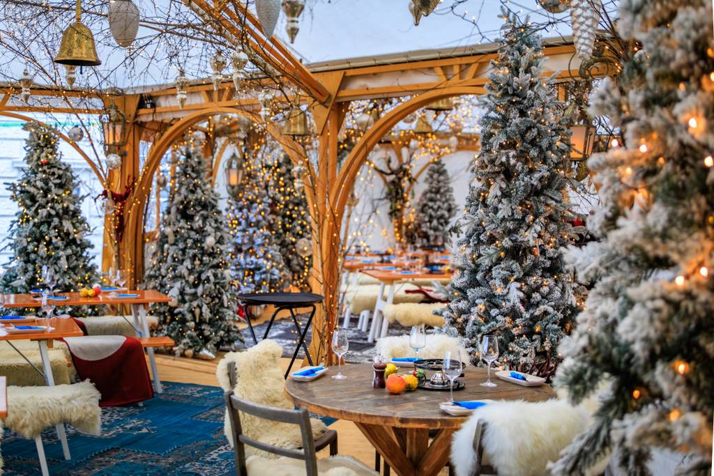 Gemütliches Restaurantinterieur im winterlichen Ambiente mit Holzbögen, schneebedeckten Weihnachtsbäumen, gedeckten Tischen mit Geschirr und warmem Licht.