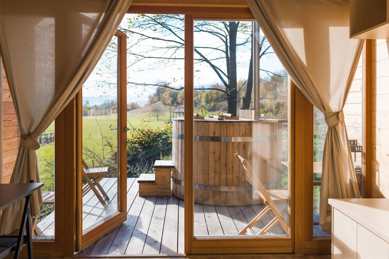 Porte a vetri aperte su una terrazza in legno con vasca idromassaggio esterna, sedie a sdraio e vista sulla campagna in autunno