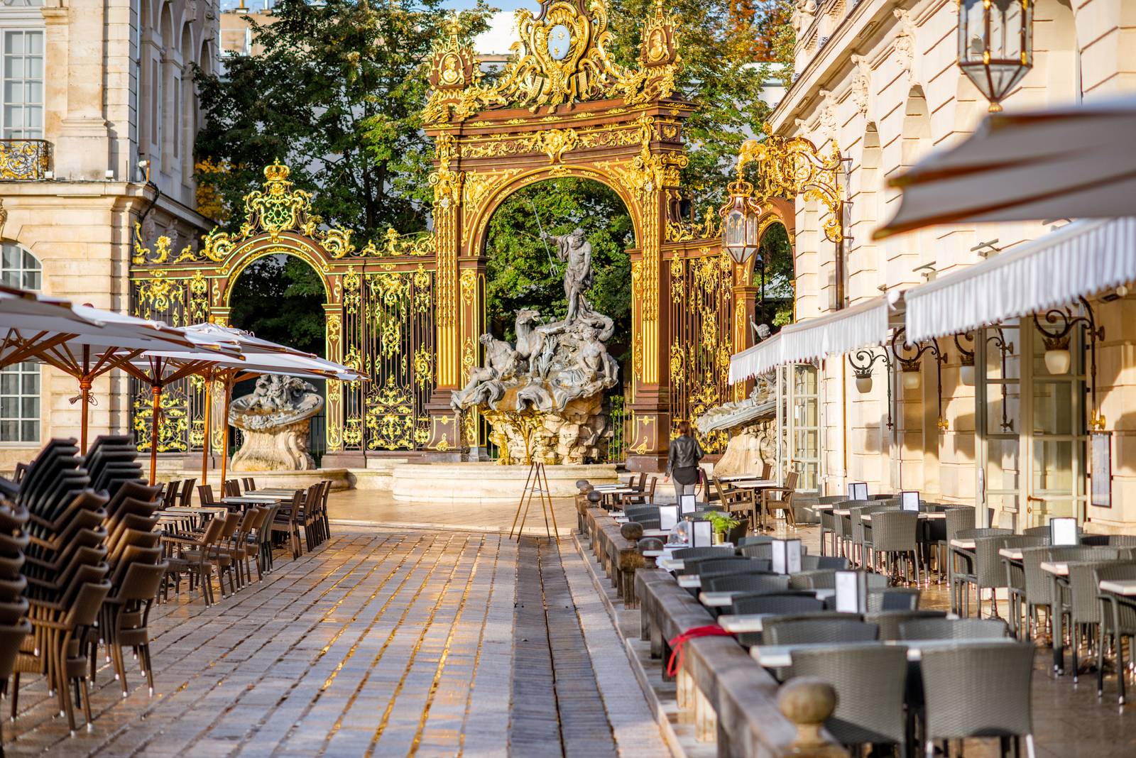 Outdoor cafe with empty tables and stacked chairs, facing an ornate golden gate and fountain, under a clear sky.