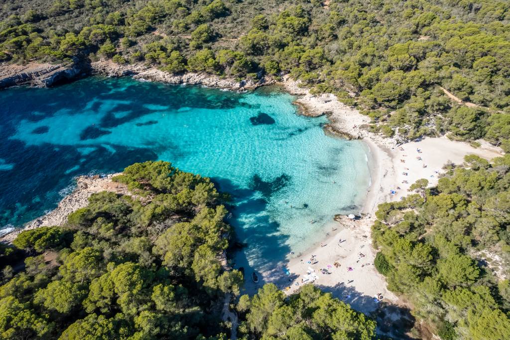 Vista aerea di Cala Turqueta, una caletta isolata dalle acque turchesi circondata da un fitto bosco verde, con bagnanti visibili sulla riva di sabbia.