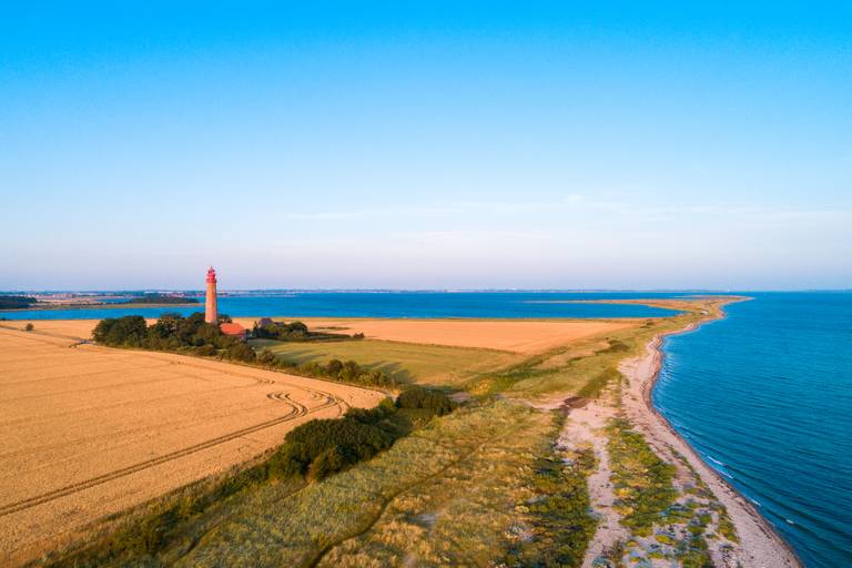 Luftaufnahme eines roten Leuchtturms an der Küste, umgeben von goldenen Feldern und blauem Meer unter klarem Himmel.
