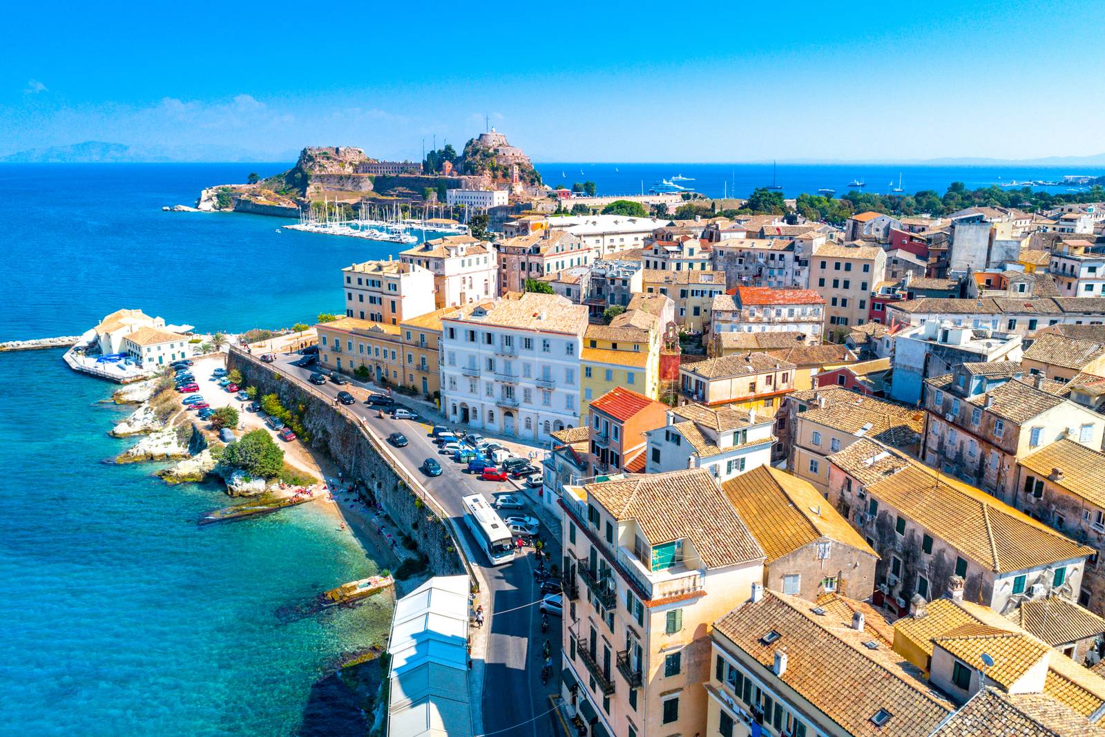 Aerial view of Corfu city with colorful buildings, a road along the shoreline, and a fortress on a hill overlooking the sea.
