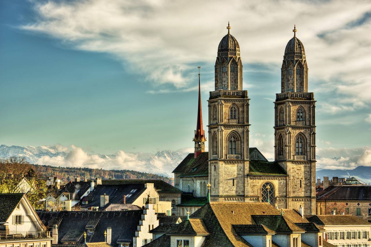 Eine historische Kathedrale mit Doppeltürmen ragt über die Dächer der Stadt empor, vor der Kulisse von Bergen und einem teils bewölkten Himmel.