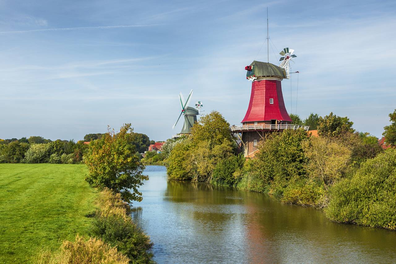 Zwei traditionelle Windmühlen an einem Fluss, umgeben von üppigem Grün und einer grasbewachsenen Wiese unter einem klaren blauen Himmel.