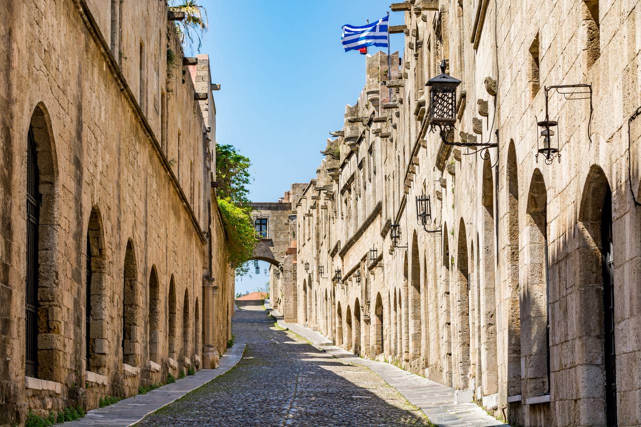 Eine Kopfsteinpflasterstraße, gesäumt von historischen Steingebäuden und gewölbten Türöffnungen, mit griechischen Flaggen und einer Laterne, unter klarem blauem Himmel