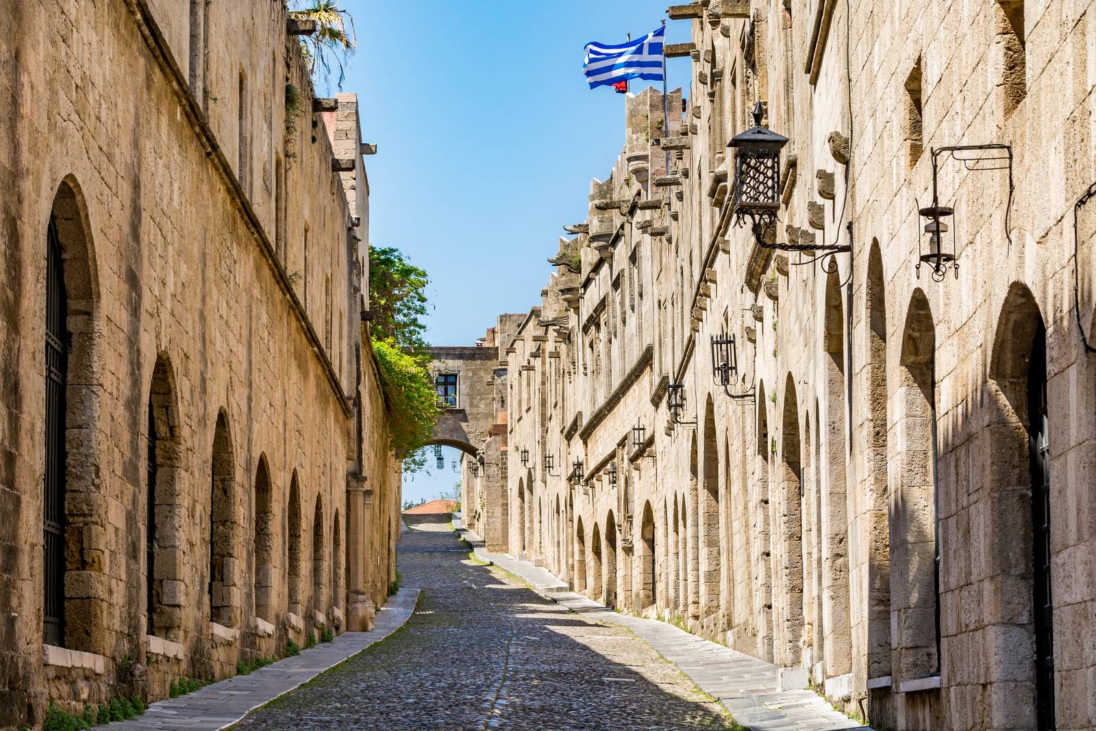 Eine Kopfsteinpflasterstraße, gesäumt von historischen Steingebäuden und gewölbten Türöffnungen, mit griechischen Flaggen und einer Laterne, unter klarem blauem Himmel