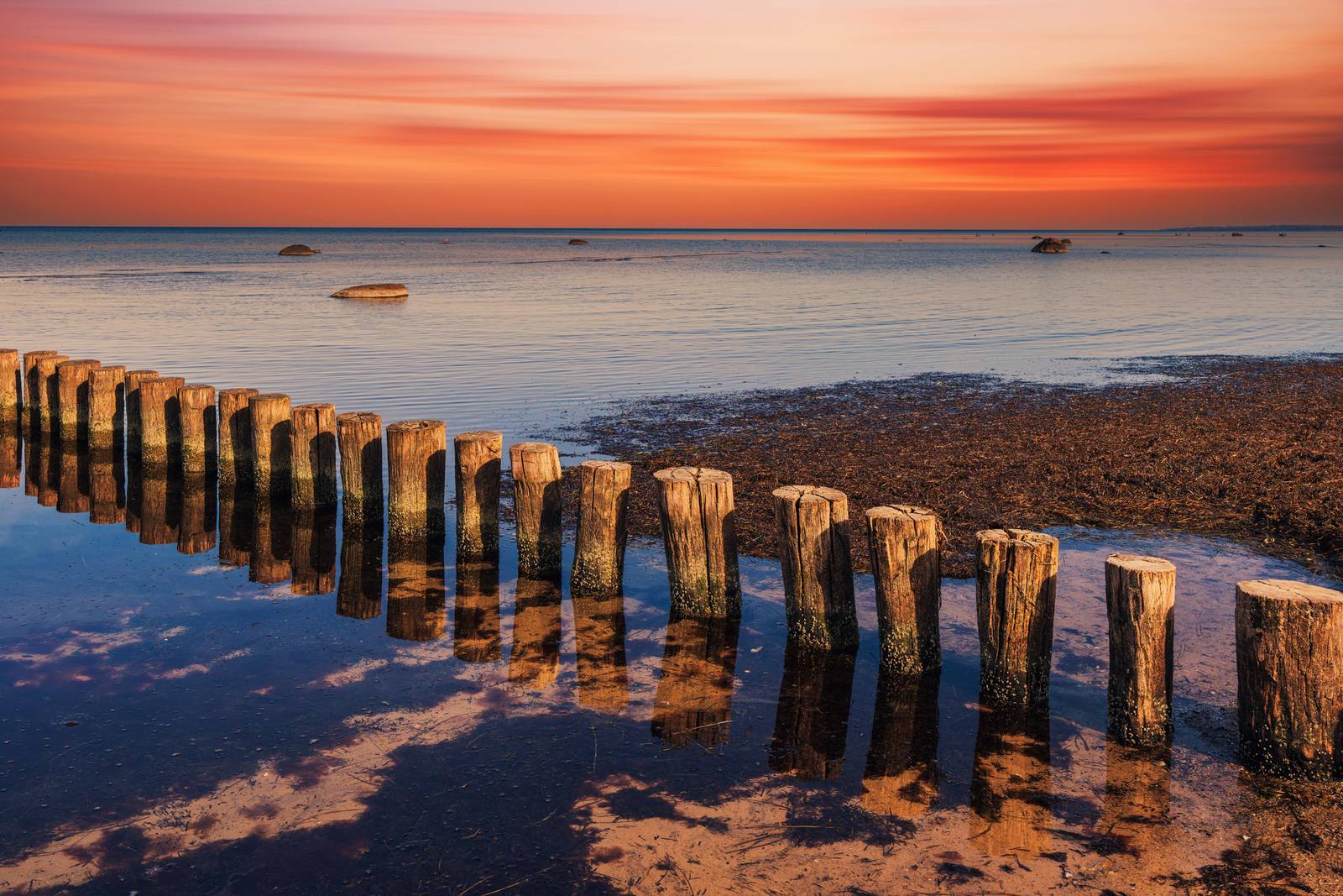 Holzpfähle im ruhigen Wasser bei Sonnenuntergang, mit einem leuchtend orangefarbenen Himmel und dessen Spiegelung auf der Wasseroberfläche.