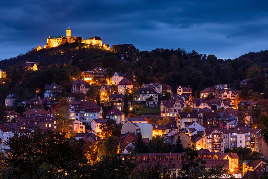 Illuminated castle on a hilltop overlooking a densely packed hillside town with warm streetlights glowing at dusk under a dark blue sky.