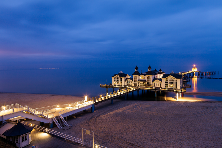 Dämmerungsblick auf einen beleuchteten Pier mit Pavillongebäuden, der sich über das ruhige Meer erstreckt, leerer Sandstrand.
