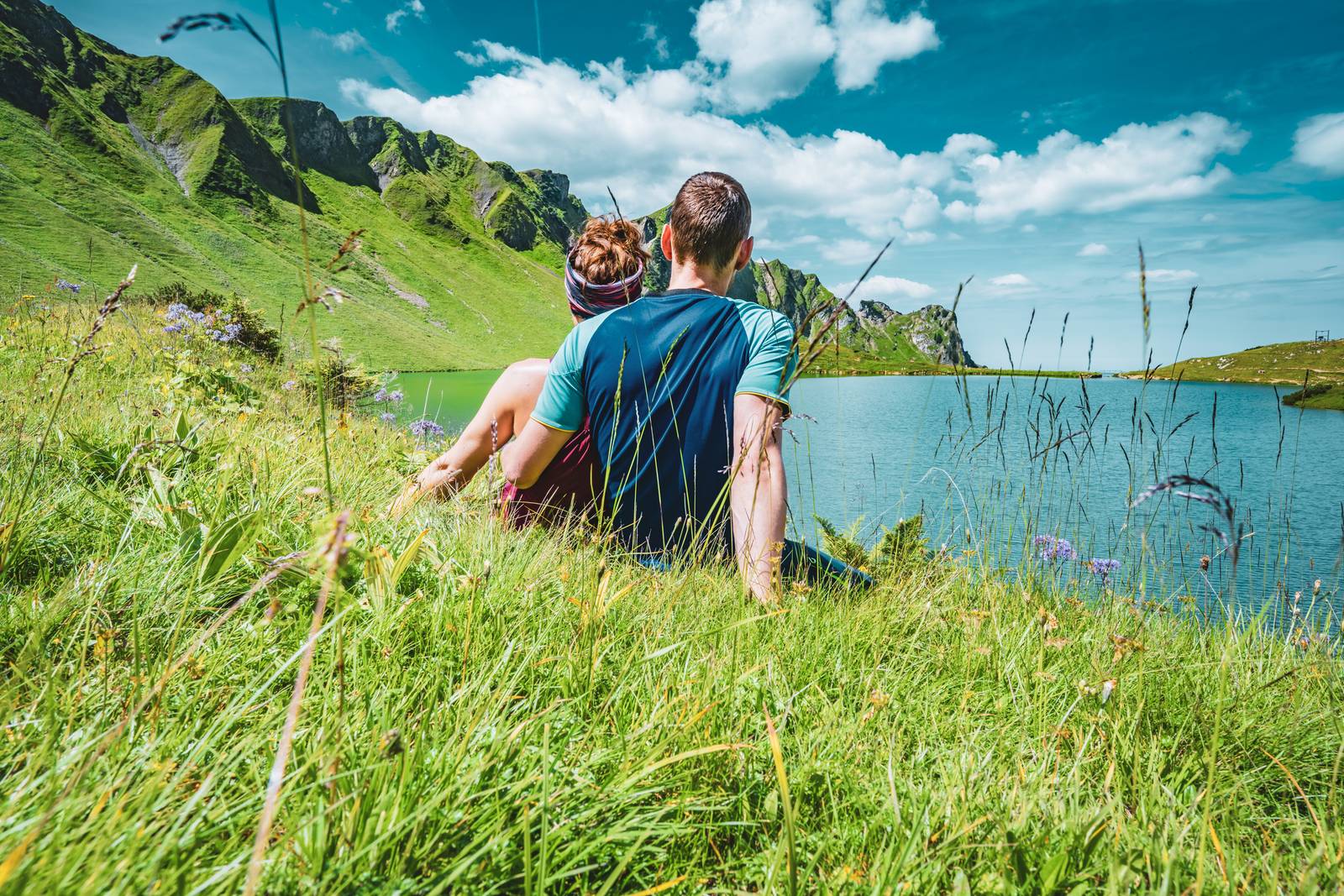 Ein Paar sitzt auf üppigem grünen Gras und blickt auf einen ruhigen See und die Berge, unter einem strahlend blauen Himmel mit vereinzelten Wolken.