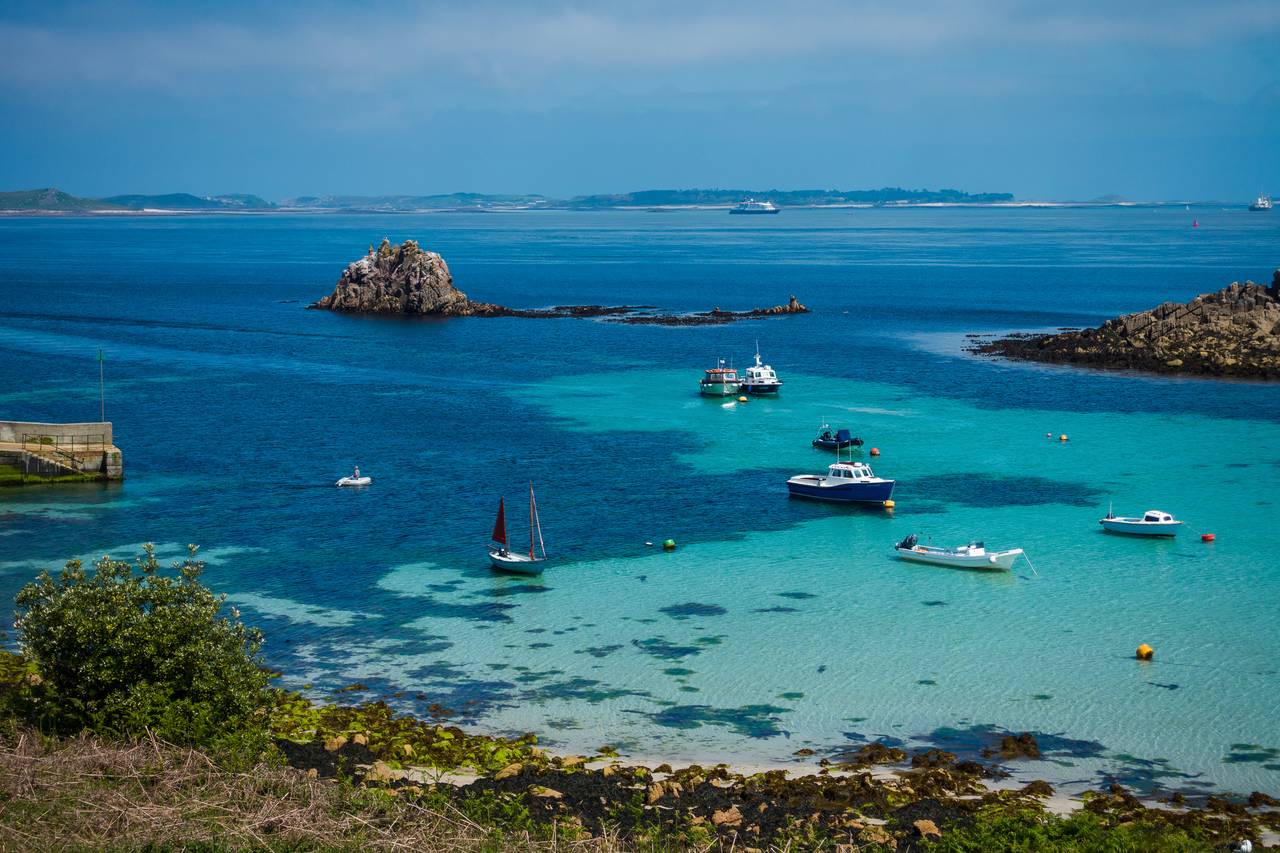 Boats anchored in a clear turquoise bay, surrounded by rocky islets and distant land, under a partly cloudy sky.