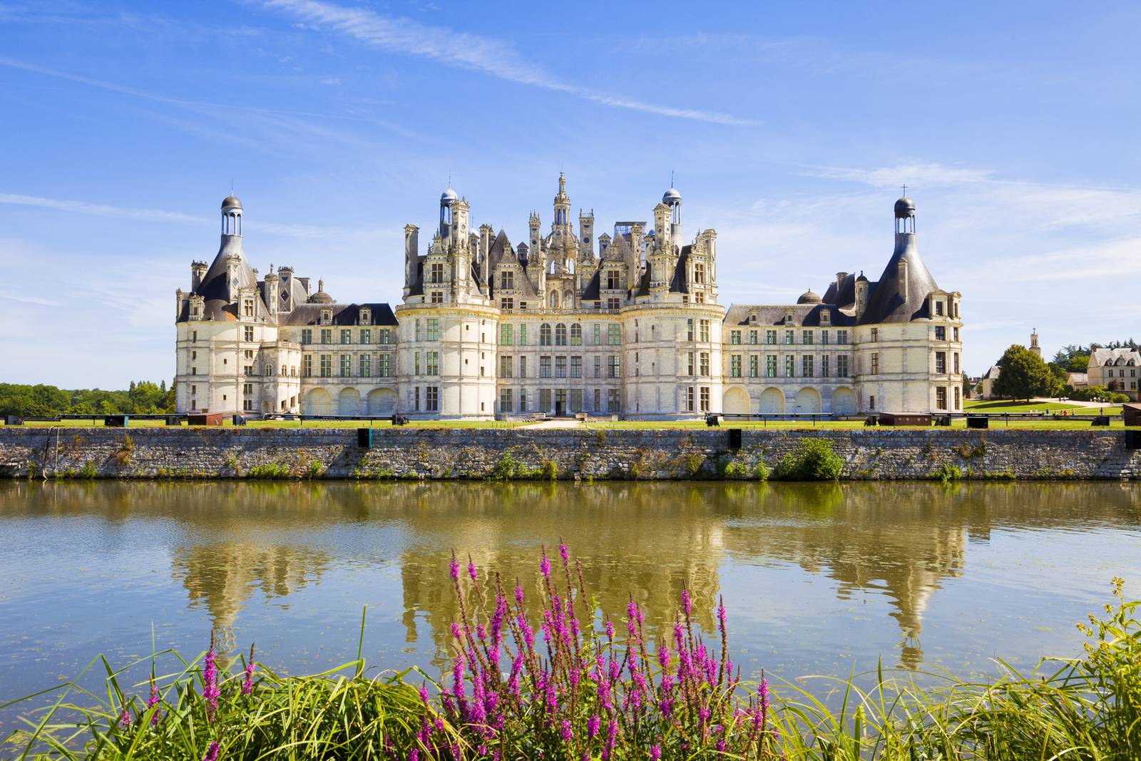 The Château de Chambord with its ornate towers and spires, reflected in a calm river, under a clear blue sky.
