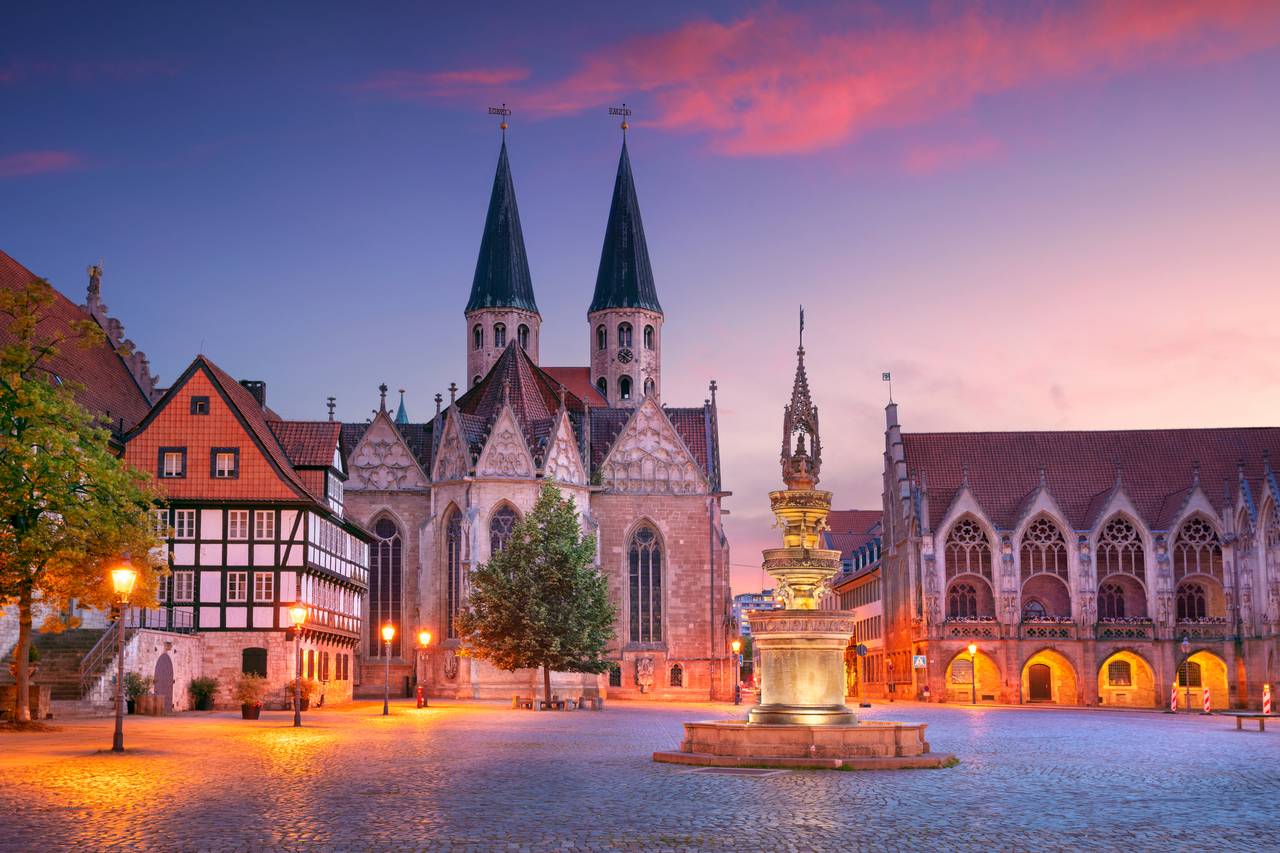 Ein historischer europäischer Platz bei Sonnenuntergang mit einer Kirche mit Doppeltürmen, einem dekorativen Brunnen und Fachwerkhäusern unter einem farbenfrohen Himmel
