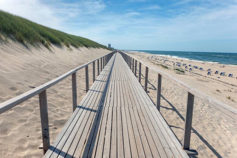 Ein hölzerner Steg führt durch sandige Dünen zu einem entfernten Strand, darüber ein blauer Himmel mit vereinzelten Wolken.
